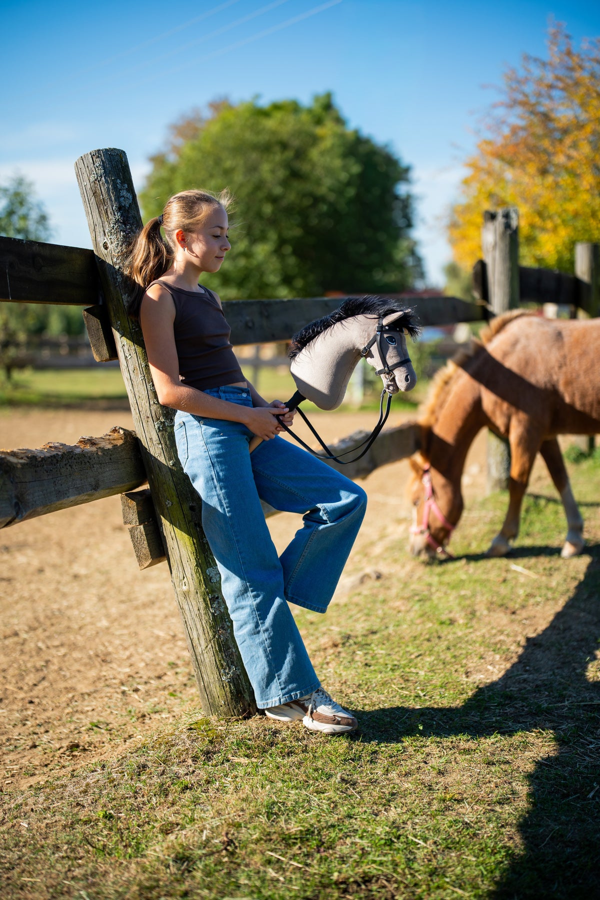 Meisje leunt in de herfstzon tegen een weidehek met Hobby Horse Thor in zwart hoofdstel, erachter graast een vospony