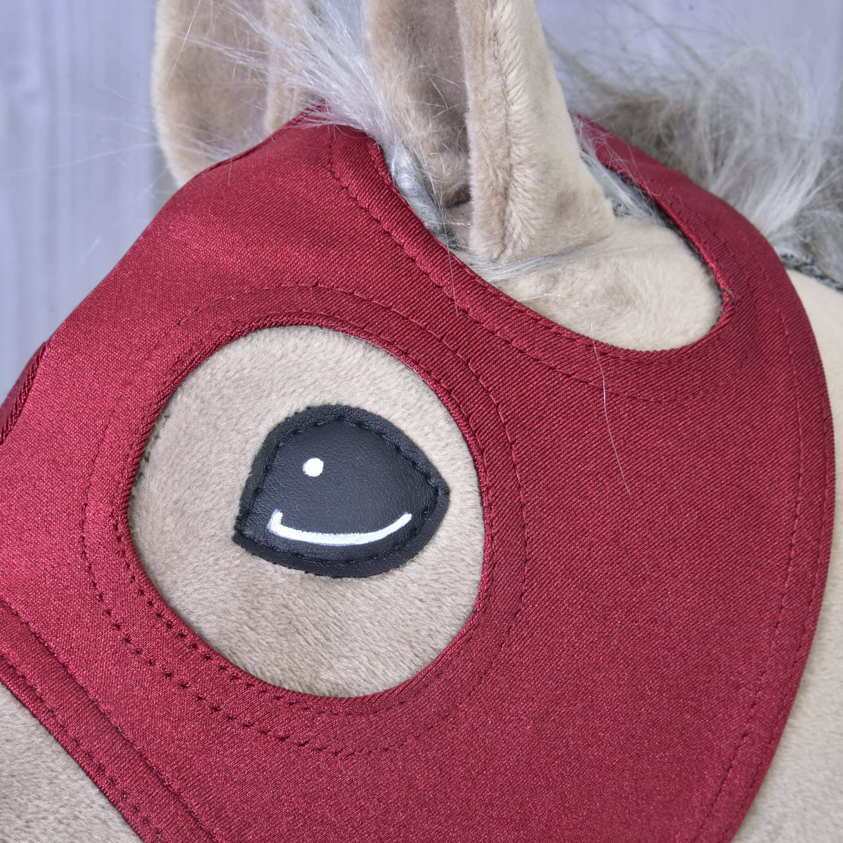 Oog en oor van een beige Hobby Horse omlijst door het bordeauxrode masker met dubbele stiksels rond de openingen en grijzige manen