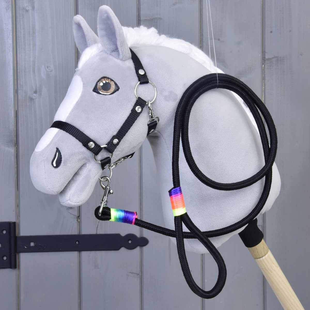 Grijze pluche Hobby Horse met zwart halster, het zwarte halstertouw met regenboogwikkels onder de kin vastgeklikt en ernaast opgerold