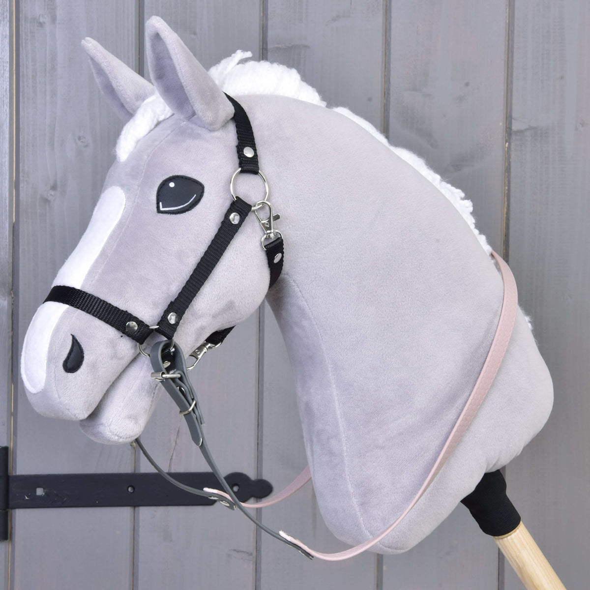 Hobby Horse van grijs pluche met zwarte halster en gerubberde teugels in donkergrijs en lichtroze, vastgegespt aan de ringen