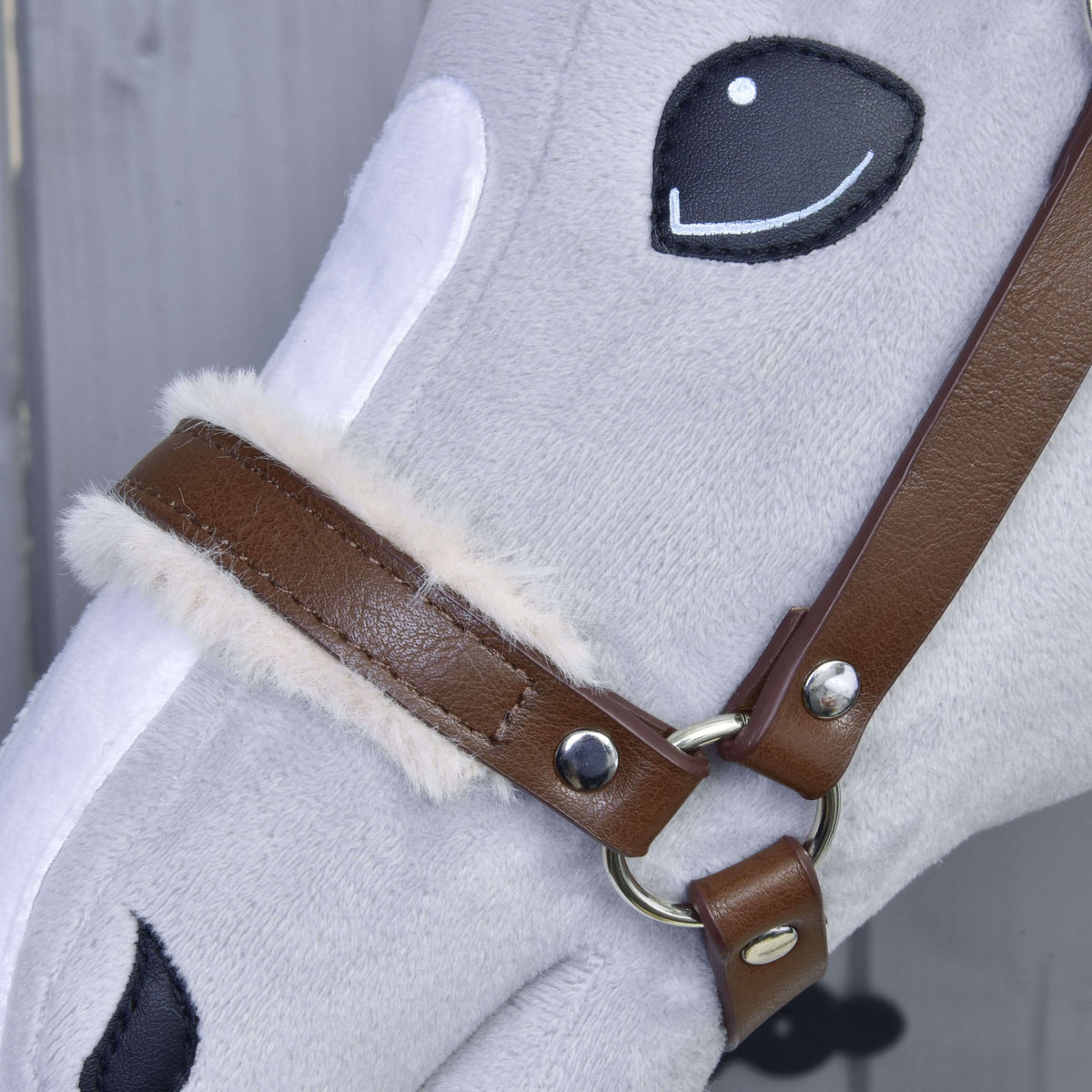 Close-up van de bruine neusriem van kunstleer met zachte crèmekleurige bontrand, zilveren klinknagels en ring op een grijze Hobby Horse