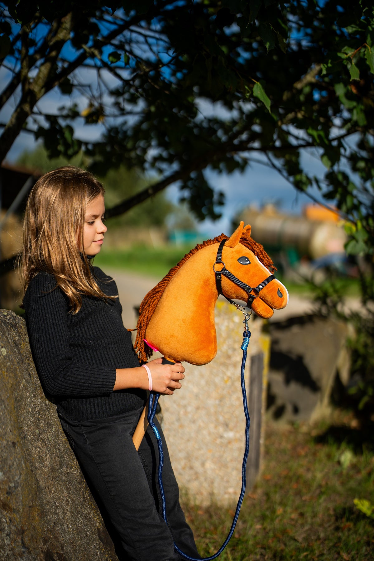 Fillette en pull noir adossée à un bloc de pierre sous un arbre, tenant Hobby Horse Wendy avec une longe bleu marine