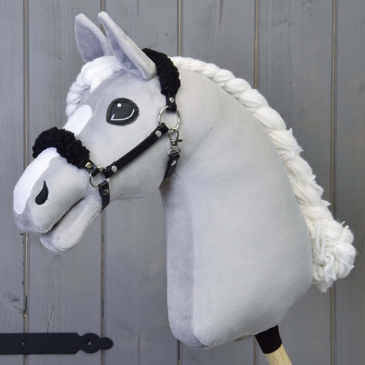 Grijze pluche Hobby Horse met gevlochten witte manen en de zwarte halster met zwart bont voor een grijze houten wand