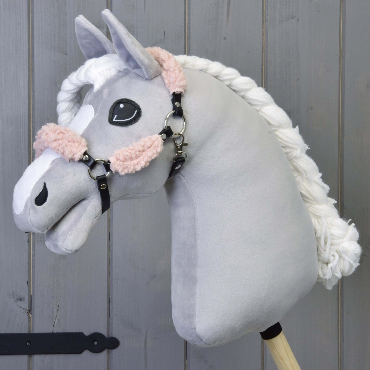 Hobby Horse gris en peluche à crinière blanche tressée, avec le licol noir à fourrure vieux rose, devant un mur en bois gris