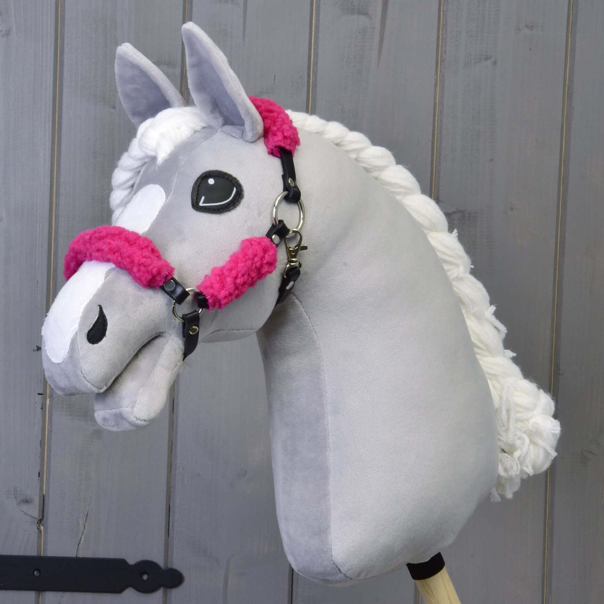 Hobby Horse gris en peluche à crinière blanche tressée, avec le licol noir à fourrure rose complète, devant un mur en bois gris