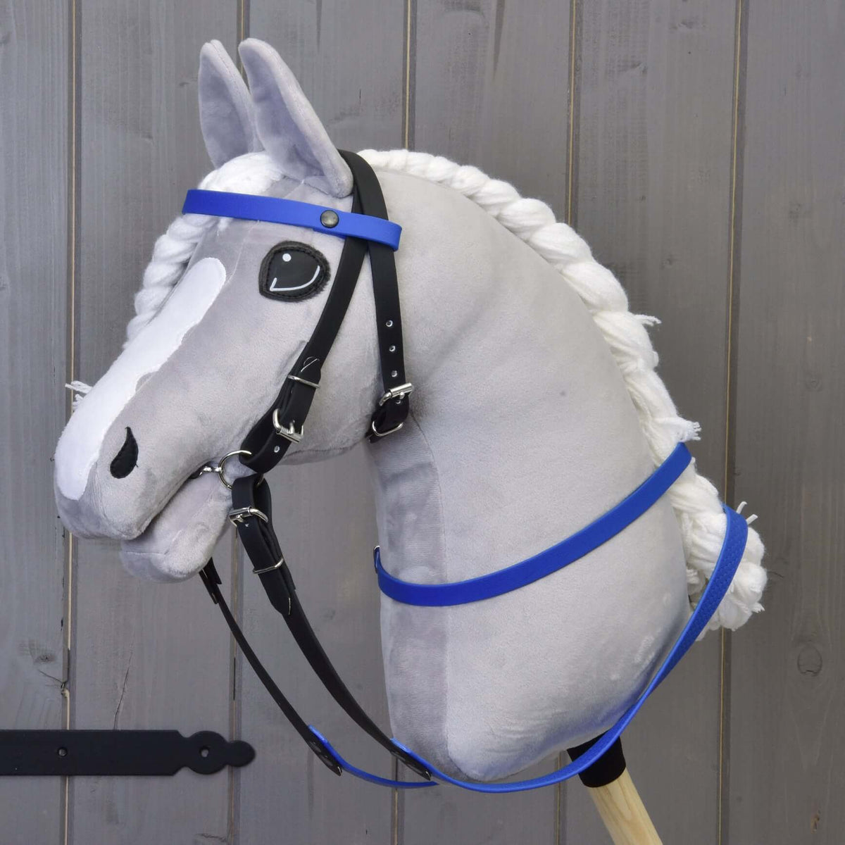 Grijze pluche Hobby Horse met zwart hoofdstel, blauwe hoofdband en de blauwe gerubberde teugels voor een grijze houten wand