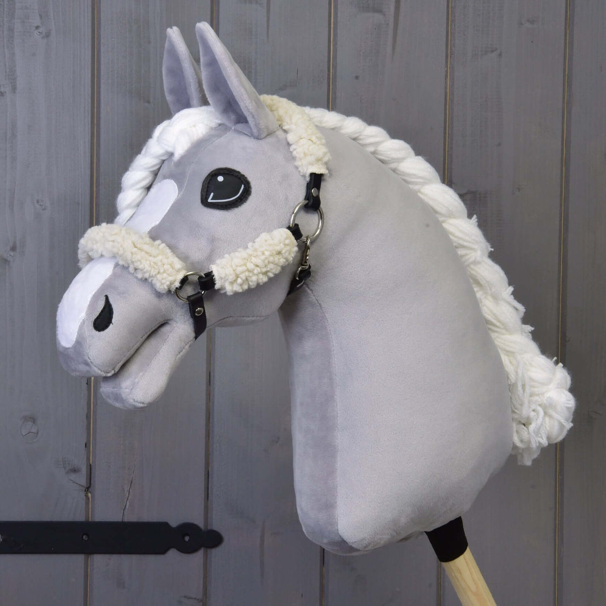 Hobby Horse gris en peluche à crinière blanche tressée, avec le licol noir à fourrure crème complète, devant un mur en bois