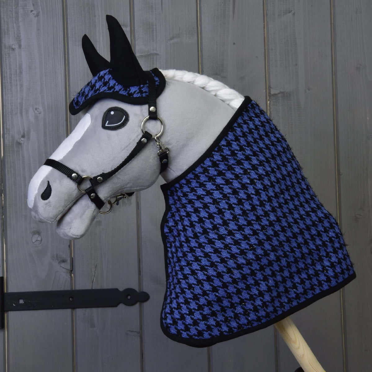 Grey plush Hobby Horse in the blue houndstooth blanket and ear bonnet with a black halter in front of a wooden wall