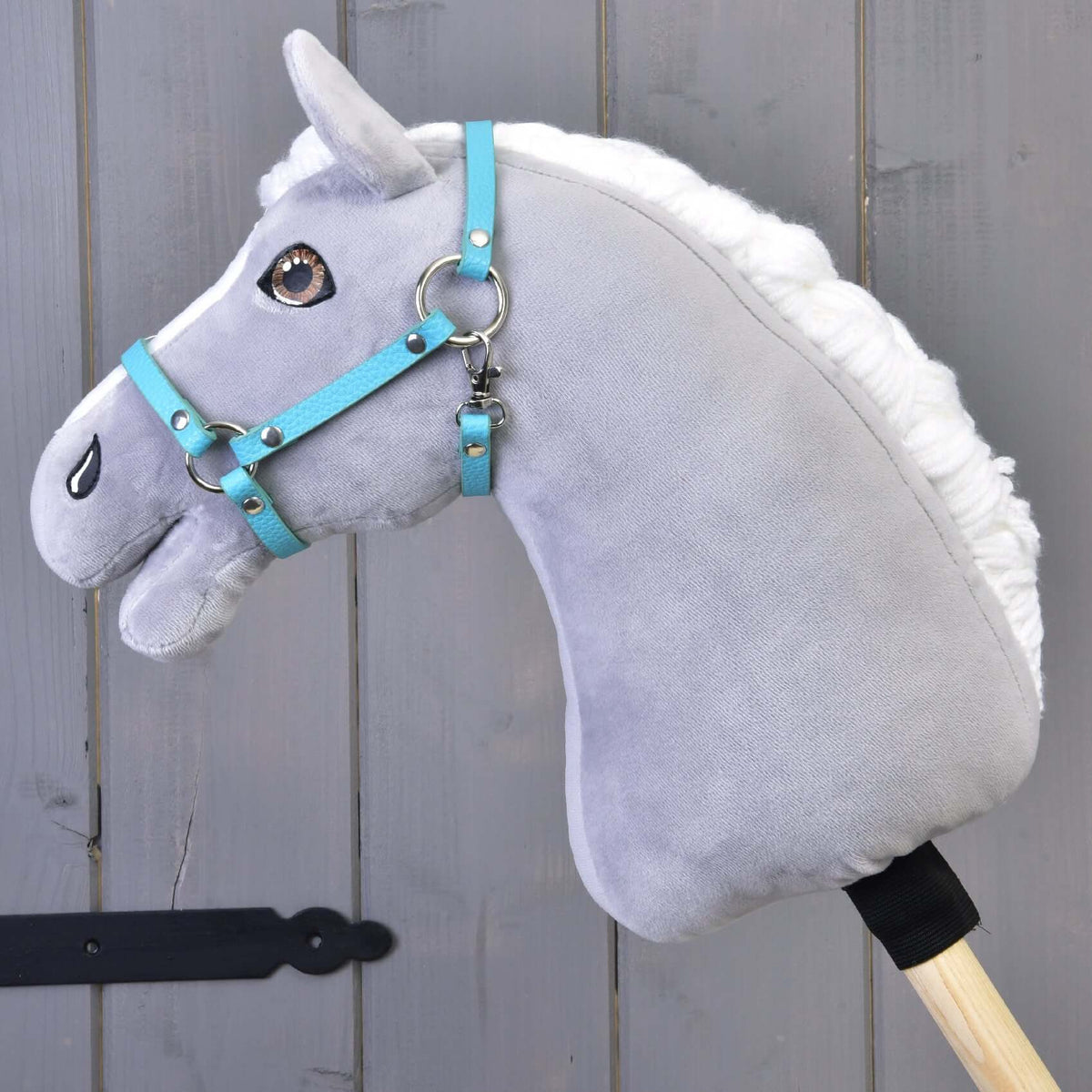 Een grijze pluche Hobby Horse met witte manen en bruine ogen draagt de turquoise halster van kunstleer voor een grijze houten wand